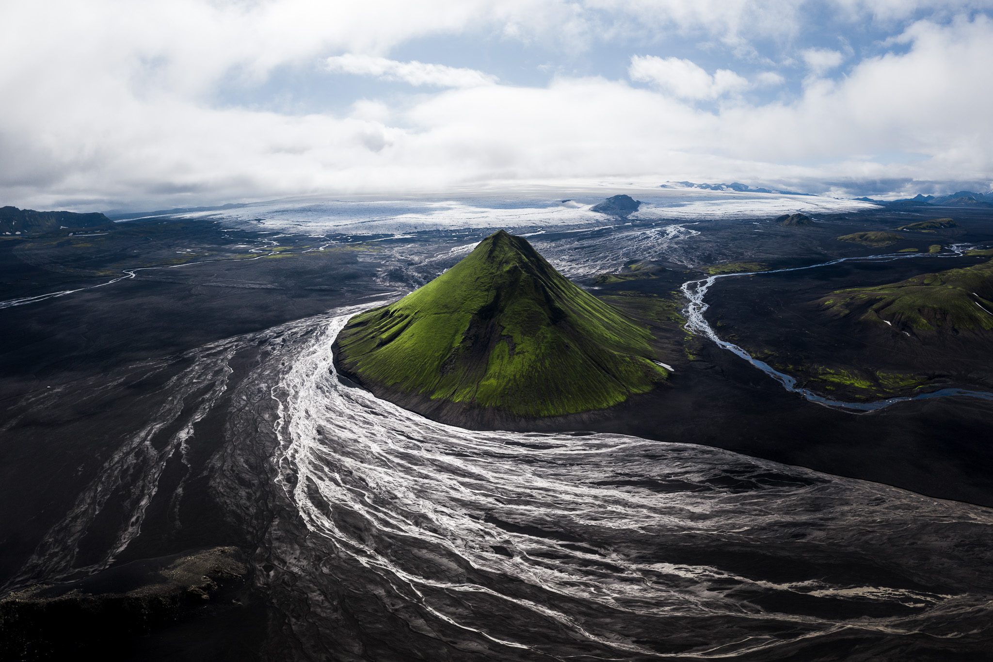 Aerial of Mælifell green pyramid mountain ringed by braided black sand streams below a glacier.
