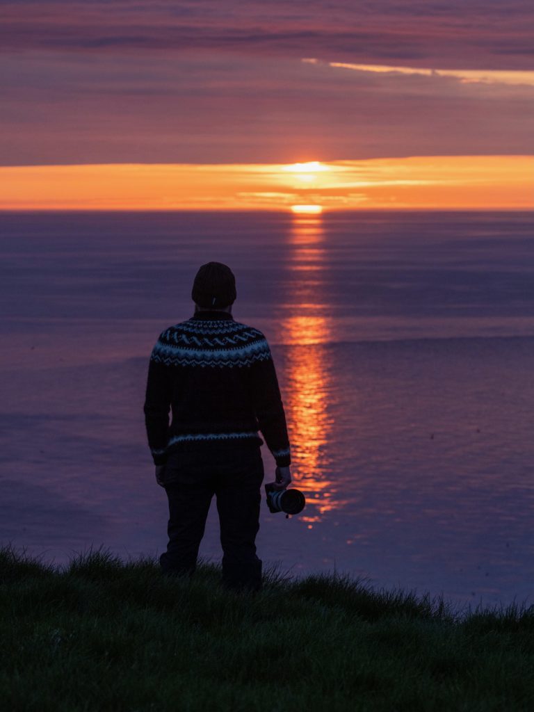 Photographer in a wool jumper watching the midnight sun set over the sea from a grassy clifftop.