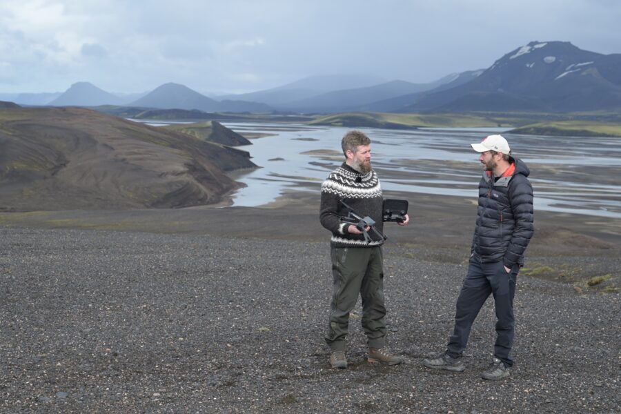 Jeroen holding a drone controller in conversation with Nigel Danson beside a braided glacial river in the Highlands