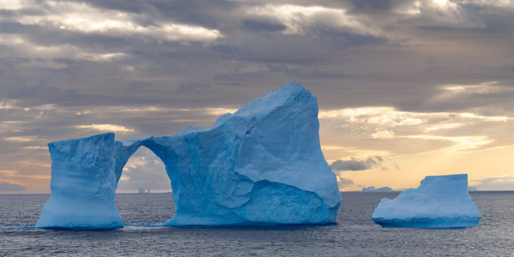 Iceberg with a natural arch under a soft pink sunset sky in Antarctic waters.