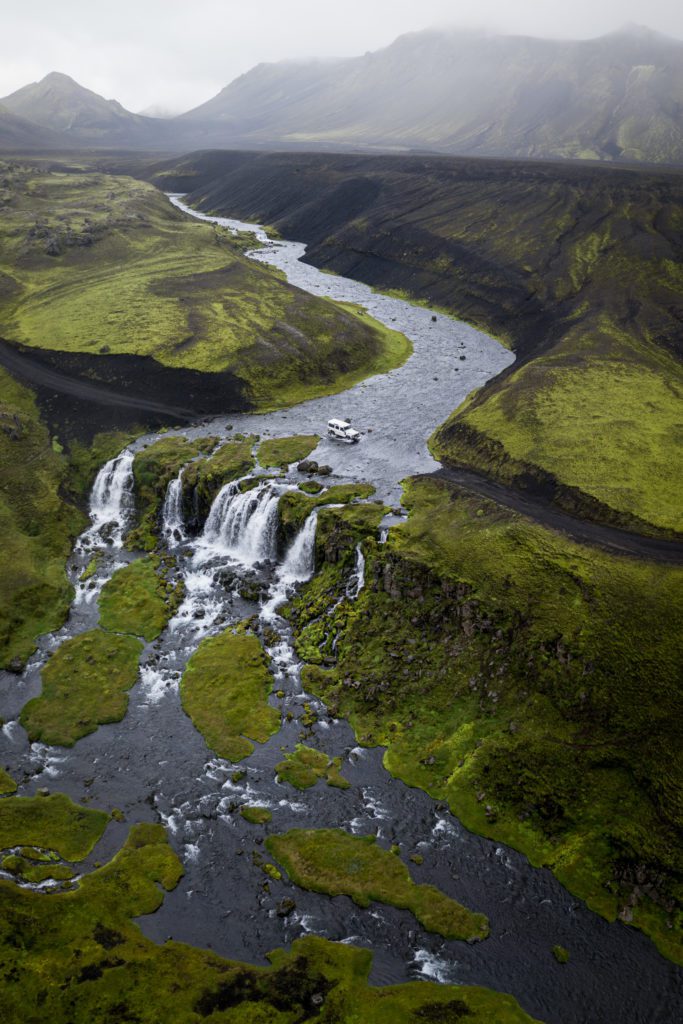Aerial drone shot of cascading waterfalls in a moss-covered Icelandic Highlands valley with a 4x4 parked above.