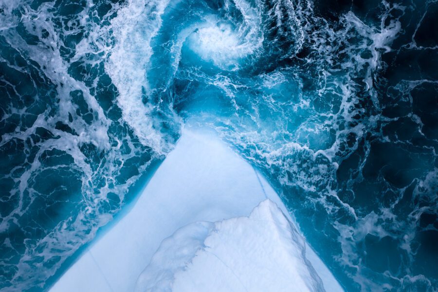 Top-down drone view of a whirlpool swirling next to the white edge of an iceberg in Greenland.